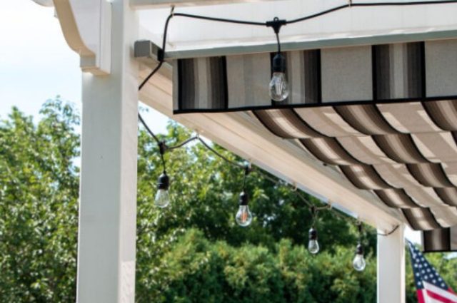 close up of white vinyl pergola top and one post with lights around the top section and a striped canopy threaded under the roof for maximum shade