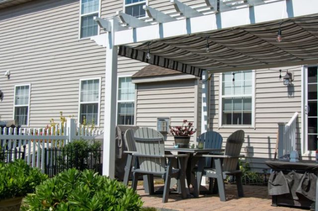 gray patio chairs and table under a detached white vinyl pergola next to green bushes