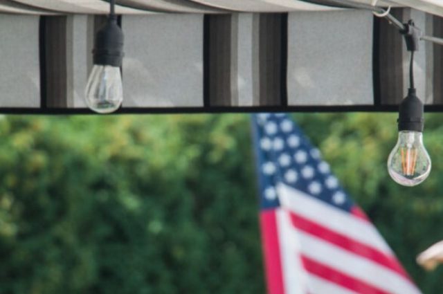 close up of a striped canopy underneath the slats of a pergola and a shot of the american flag in the distance