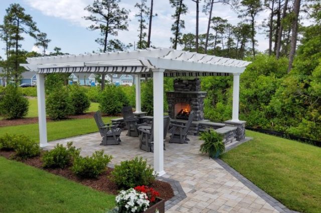 white detached pergola beside a manicured lawn with a stone fireplace and polywood furniture under it