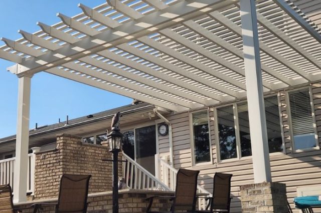 white vinyl pergola that is attached to the back of a home with posts in the front and over an stone outdoor fireplace