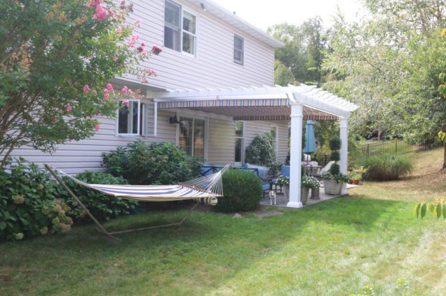 White pergola with striped canopy over back porch, attached to beige house, with blue outdoor furniture and plants.