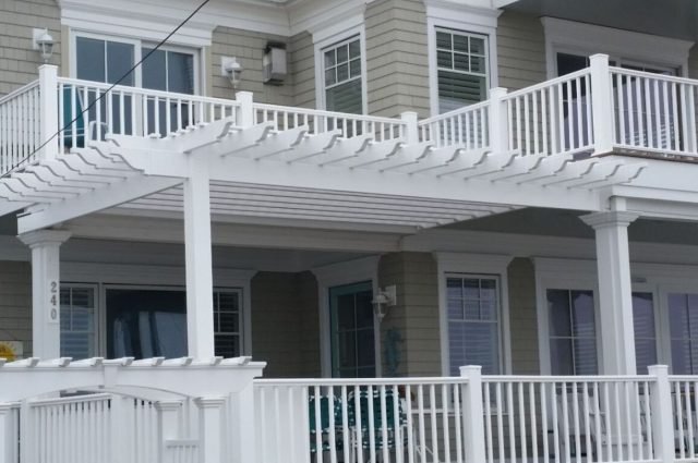 A beach house with a white vinyl pergola attached over the patio, featuring matching white railings on the upper deck and front porch.