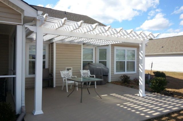 White pergola over concrete patio attached to beige house, featuring outdoor chairs and a grill for a cozy seating space.