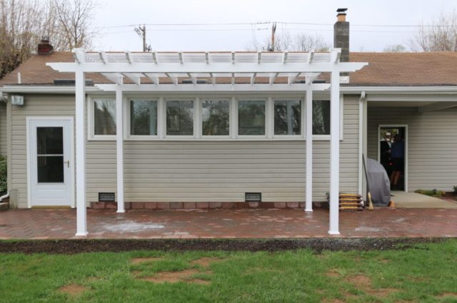 White pergola with posts over a brick patio, attached to beige house, providing a shaded outdoor space.