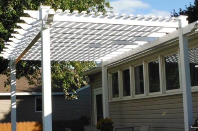 A white pergola in a backyard, providing shade over a patio with a table and chairs. The structure is adjacent to a house with large windows and is set against a backdrop of trees and greenery.