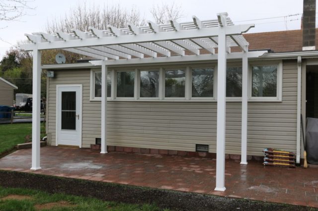 White freestanding pergola with brick flooring in a backyard, attached to a beige house, adding outdoor elegance.