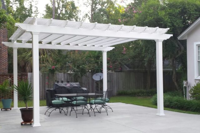 A freestanding white vinyl pergola providing shade over an outdoor dining area on a concrete patio.