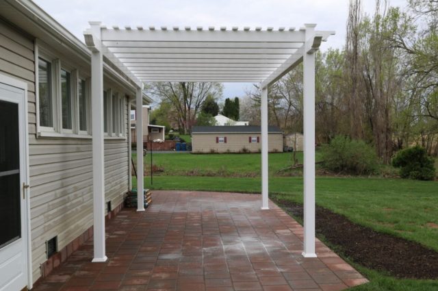 White freestanding vinyl pergola over brick patio, attached to beige house, offering a stylish outdoor area.