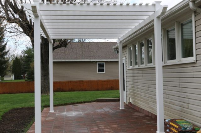 Freestanding white pergola on brick patio, attached to beige house, enhancing backyard aesthetics.