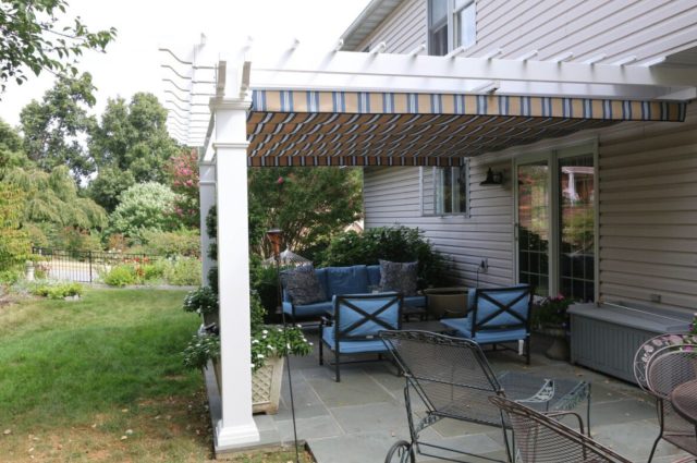 White pergola with striped canopy over garden patio, attached to beige house, surrounded by lush greenery and blue outdoor seating.