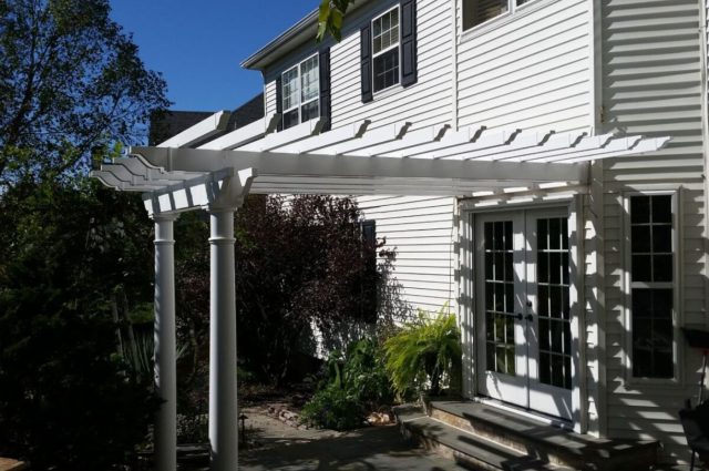 A white pergola attached to a house, creating a shaded outdoor area. The structure is supported by columns and is adjacent to a double door entrance, surrounded by greenery and plants.