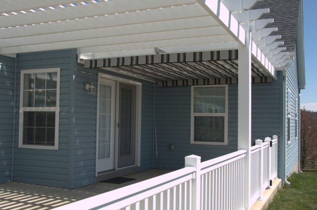 Contemporary white vinyl pergola with striped canopy, attached to a blue house with white railings.