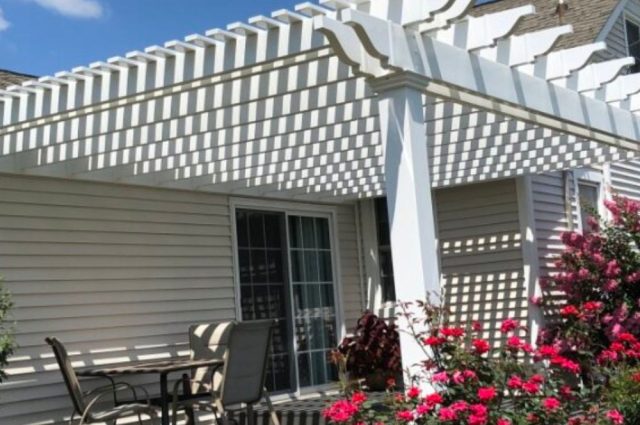 A white pergola providing shade over a patio area with outdoor furniture. The structure is surrounded by blooming pink flowers and lush green bushes, creating a cozy and inviting outdoor space.