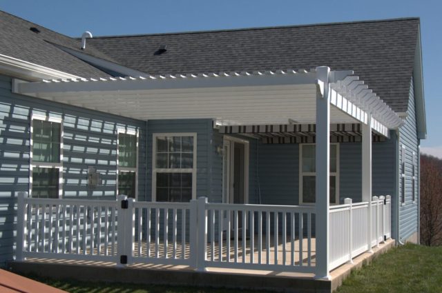 White vinyl pergola attached to blue house, featuring white railings and providing shaded outdoor space.