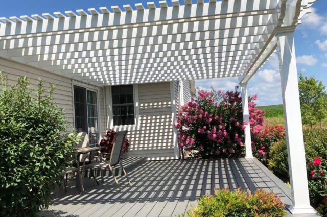 A white pergola elevated over a wooden deck, providing shade and a pleasant outdoor sitting area. The deck is surrounded by vibrant pink flowers and green plants, enhancing the beauty of the space.