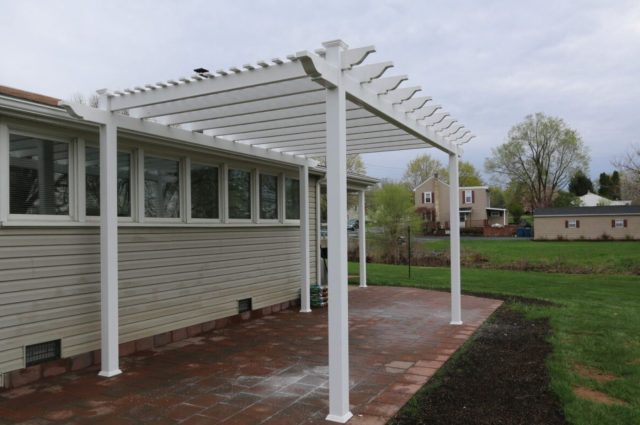 White vinyl pergola attached to beige house, overlooking backyard, with installer in the background.