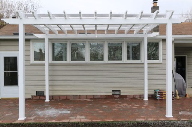 White pergola with posts over brick patio, attached to beige house, creating a shaded outdoor living space.