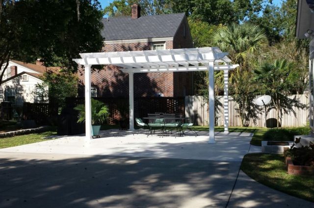 A white vinyl pergola providing shade over a concrete patio with outdoor furniture in a backyard.