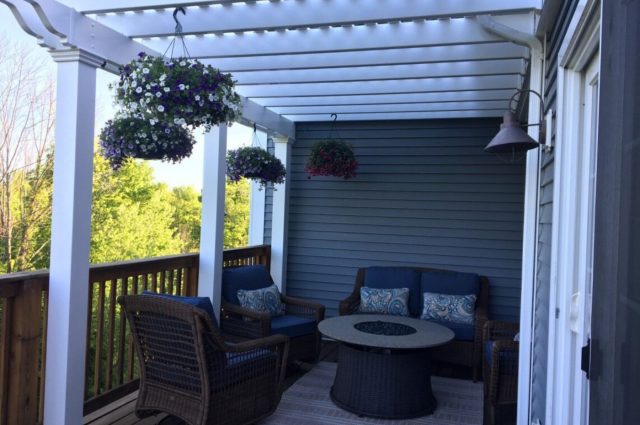 White pergola over deck attached to blue two-story house, with wicker furniture, a fire pit, and hanging flower baskets.