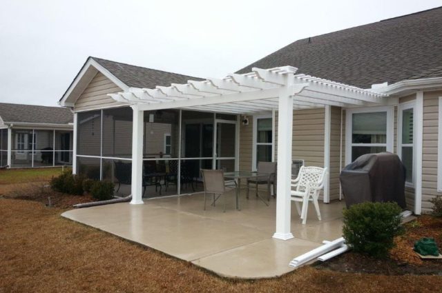 White pergola attached to beige house over patio with outdoor chairs and a grill, creating a shaded relaxation spot.