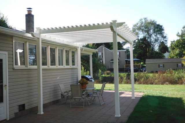 White vinyl pergola over brick patio, attached to beige house, providing a stylish shaded area.