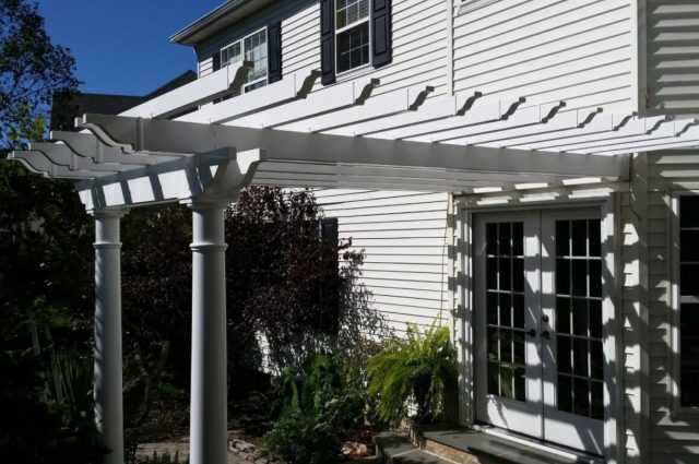 A white vinyl pergola over a patio, providing a shaded area with outdoor seating. The pergola is surrounded by green bushes and vibrant pink flowers, creating a serene and relaxing outdoor environment.