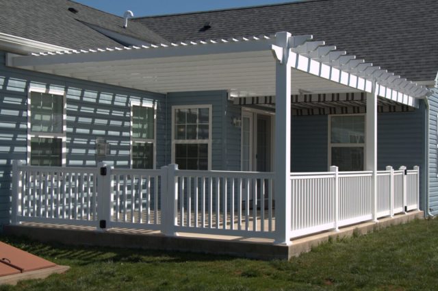 Vinyl pergola with white railings around a porch, attached to a blue house, creating a shaded outdoor area.